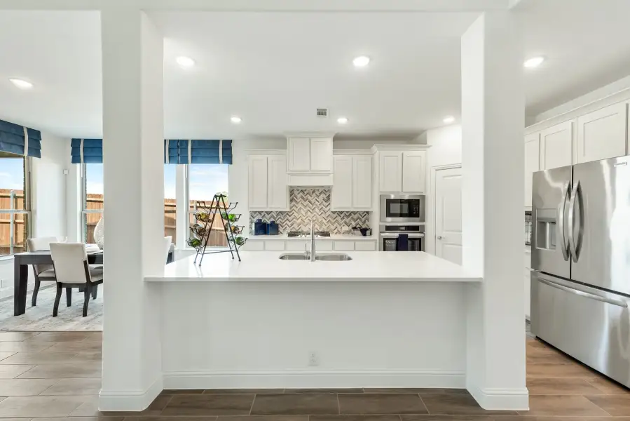 Kitchen with white cabinets, herringbone tile backsplash, stainless steel appliances, and white island with sink open to dining area.