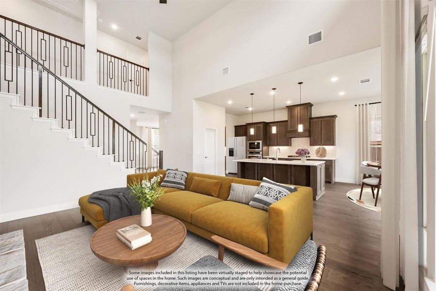 Open-concept living room with two-story ceiling, staircase, and kitchen visible in background, hardwood floors throughout.