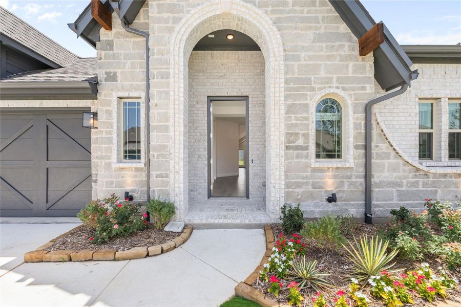 Stone home entrance with arched doorway, gray front door, and landscaped flower beds along walkway Stone home entrance with arched doorway, gray front door, and landscaped flower beds along walkway