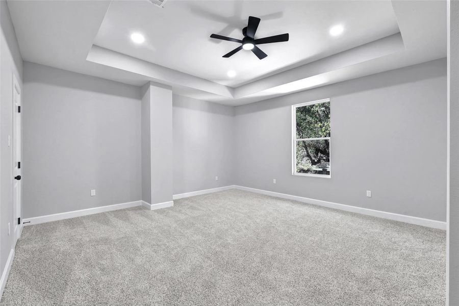This room features neutral gray walls and carpet, a tray ceiling with recessed lighting, and a modern ceiling fan. A large window provides natural light and a view of greenery.