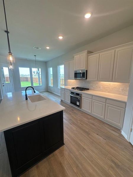 Kitchen featuring a central island with an undermount sink, light-toned countertops, wood-finish flooring, a stainless steel microwave and oven, and white cabinetry