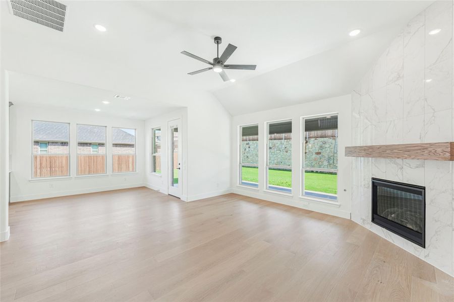 Open living room with light hardwood floors, marble tile fireplace, ceiling fan, and large windows overlooking backyard. Open living room with light hardwood floors, marble tile fireplace, ceiling fan, and large windows overlooking backyard.