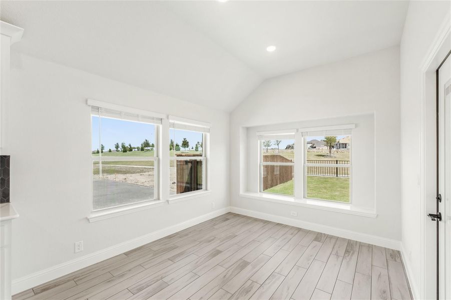Empty room with vaulted ceiling, light wood floors, and four windows overlooking backyard
