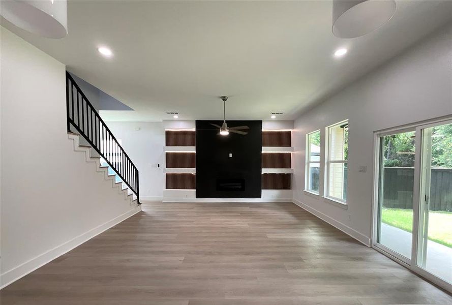 Spacious living area featuring wood-finish flooring, a modern black fireplace wall with integrated shelving and accent lighting, and a contemporary black metal staircase railing Spacious living area featuring wood-finish flooring, a modern black fireplace wall with integrated shelving and accent lighting, and a contemporary black metal staircase railing