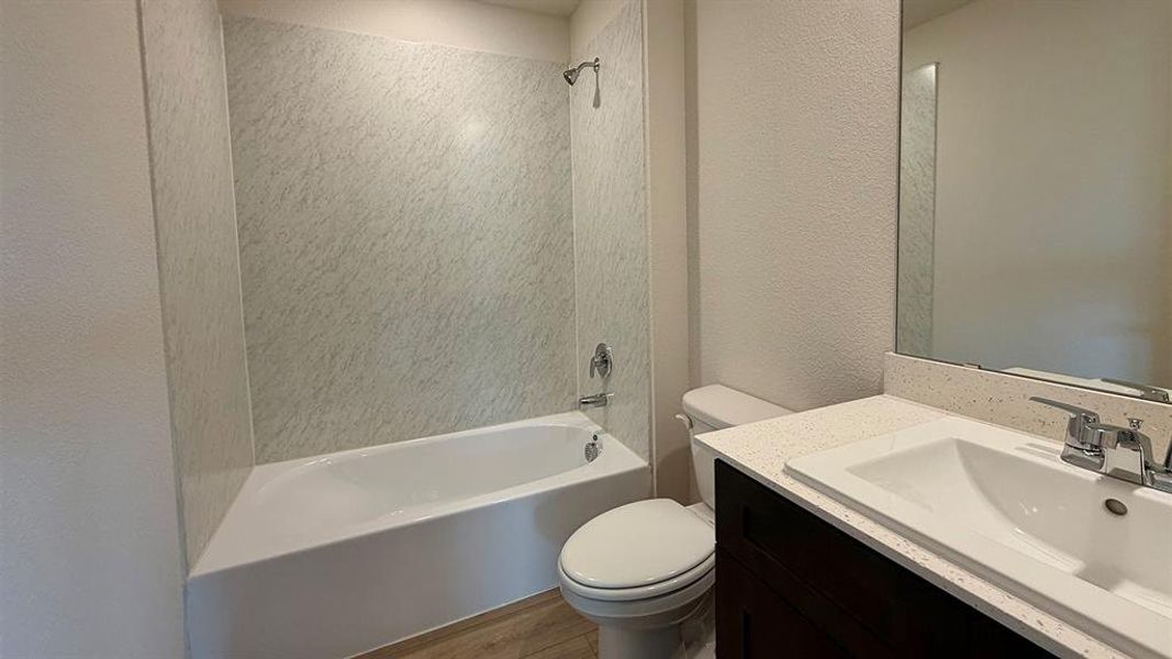 Full bathroom featuring a white bathtub with a showerhead, a patterned shower surround, a white toilet, a dark wood-finish vanity, and a light-toned countertop with an integrated sink