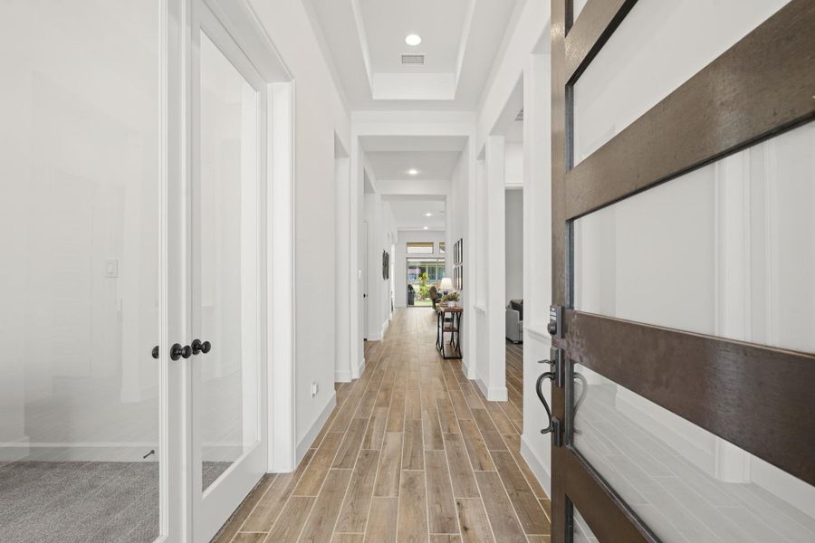 This photo showcases a modern, inviting entryway with a sleek wooden door and glass panels. The hallway features wood-look tile flooring, bright white walls, and recessed lighting, leading to an open living area.