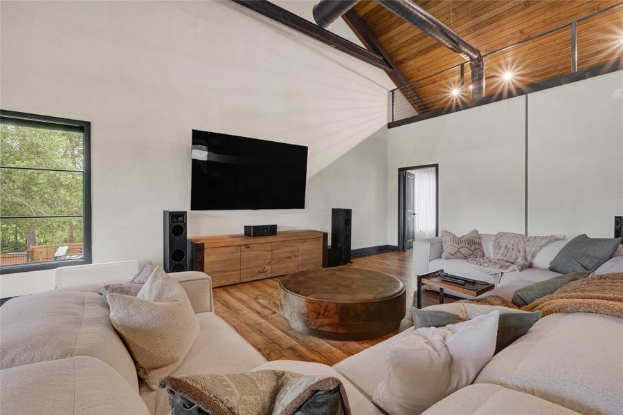 Vaulted wooden ceiling, large sectional sofa, and a wall-mounted TV. It has a warm, inviting ambiance with plenty of natural light from a large window, and modern decor elements like a sleek coffee table and contemporary speakers.