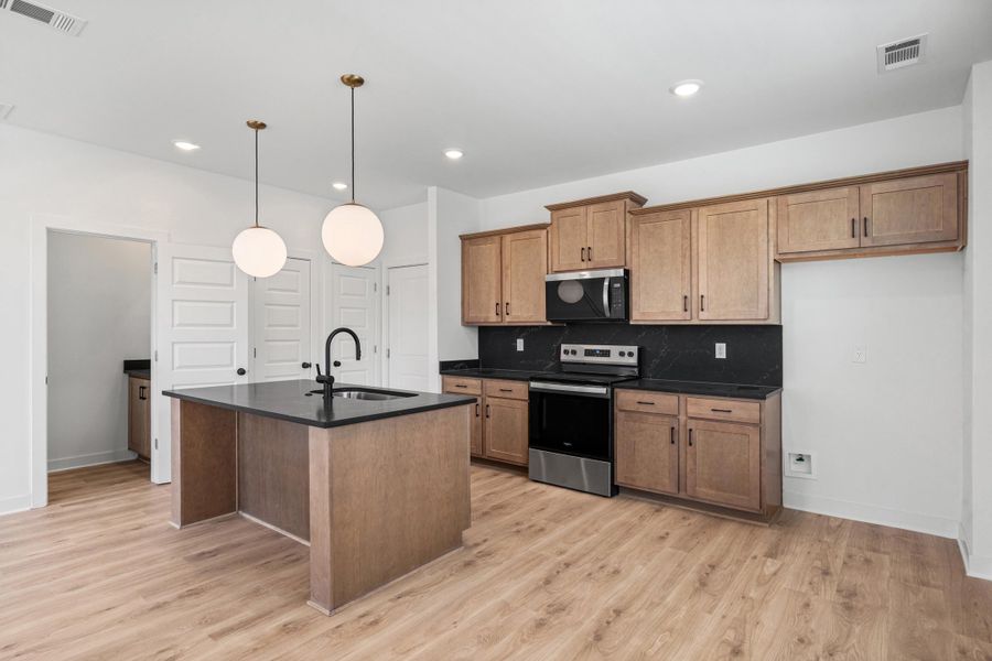Kitchen featuring wood-finish flooring, a kitchen island with a black countertop and integrated sink, pendant lighting, wood-finish cabinetry, and stainless steel appliances