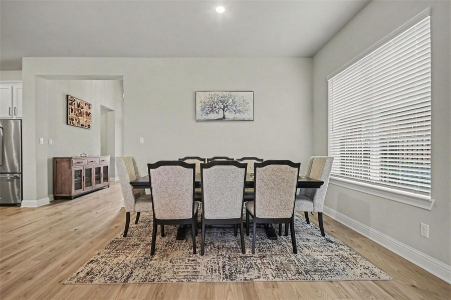This dining area features a spacious table for eight, set on a stylish rug. Large windows with blinds provide natural light, and the open layout includes a view into the kitchen with stainless steel appliances. Light wood flooring adds a warm touch. This dining area features a spacious table for eight, set on a stylish rug. Large windows with blinds provide natural light, and the open layout includes a view into the kitchen with stainless steel appliances. Light wood flooring adds a warm touch.