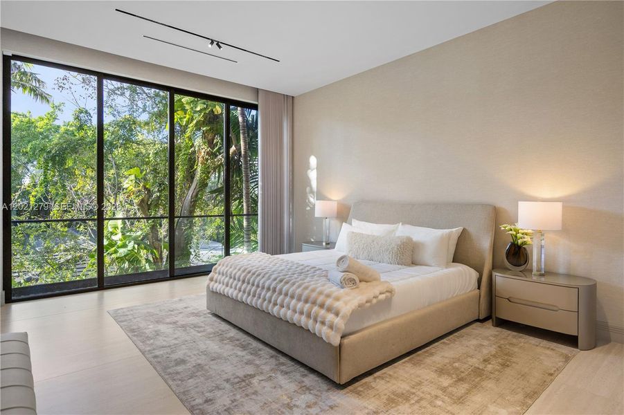 Bright en-suite bedroom with upholstered king bed, matching nightstands with table lamps, neutral textured walls, light stone floors, area rug, sheer drapery, track lighting, and floor-to-ceiling sliding glass doors with lush tropical garden views.