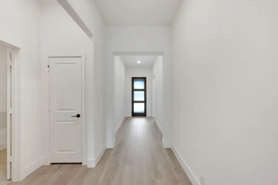 Hallway with light wood floors, white walls, and dark glass front door at the end