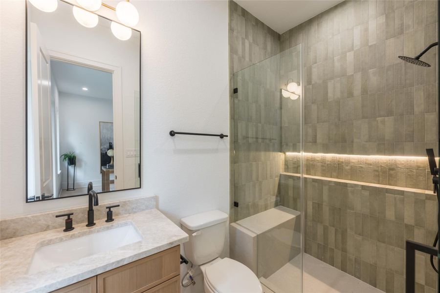 On the spec sheet: handmade zellige-inspired wall tile in a warm sage glaze, a frameless glass shower with integrated LED niche, a rainfall head paired with a wall-mount on matte-black trim, a light-oak floating vanity with undermount sink under a stone slab top, and a black-framed mirror lit by an opal-globe brass fixture.