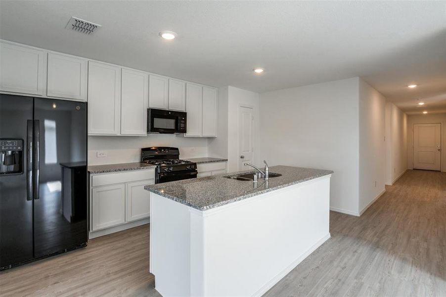 Kitchen featuring white cabinets, black appliances, a kitchen island with sink, light stone countertops, and light wood-type flooring