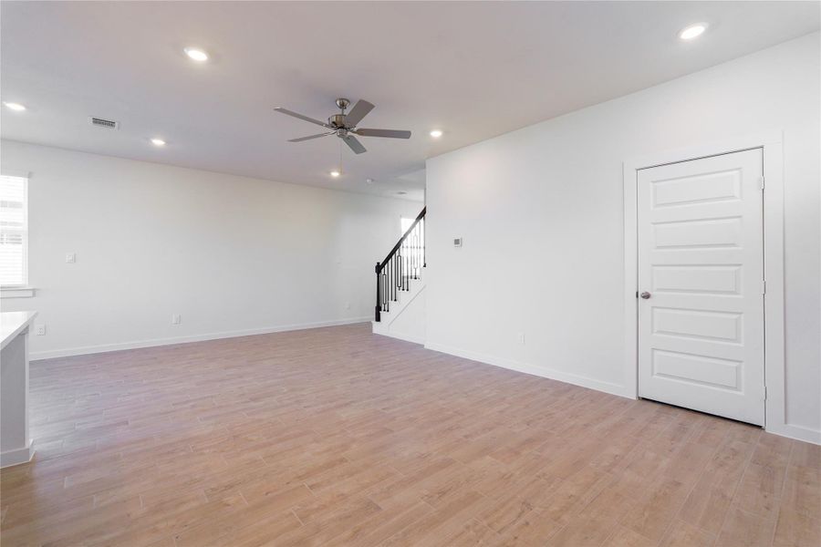 This photo shows a spacious, open-concept room with light wood flooring and neutral walls. It features recessed lighting, a ceiling fan, and a staircase with dark railings. A white door is present, enhancing the modern, clean design.