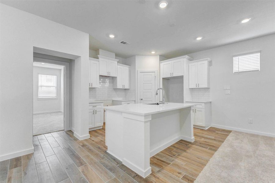 Kitchen view featuring a large white island, bright cabinetry, subway tile backsplash, recessed lighting, wood-look tile flooring, and open access to the nearby living space. The clean design offers a modern, functional workspace with room for meal prep, serving, and gathering.