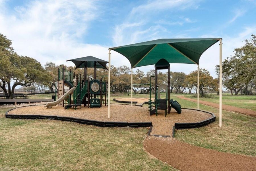 Outdoor recreational area featuring a multi-level play structure with slides and climbing elements, shaded by a dark green canopy
