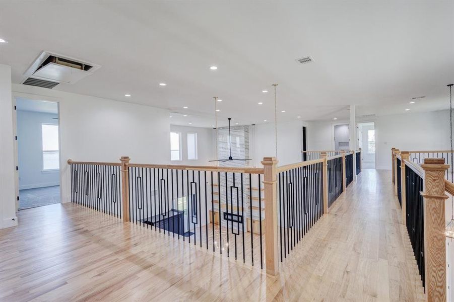 Expansive second-story landing featuring wood-finish flooring, a contemporary black metal and wood baluster railing, recessed ceiling lighting, and multiple windows providing ambient light
