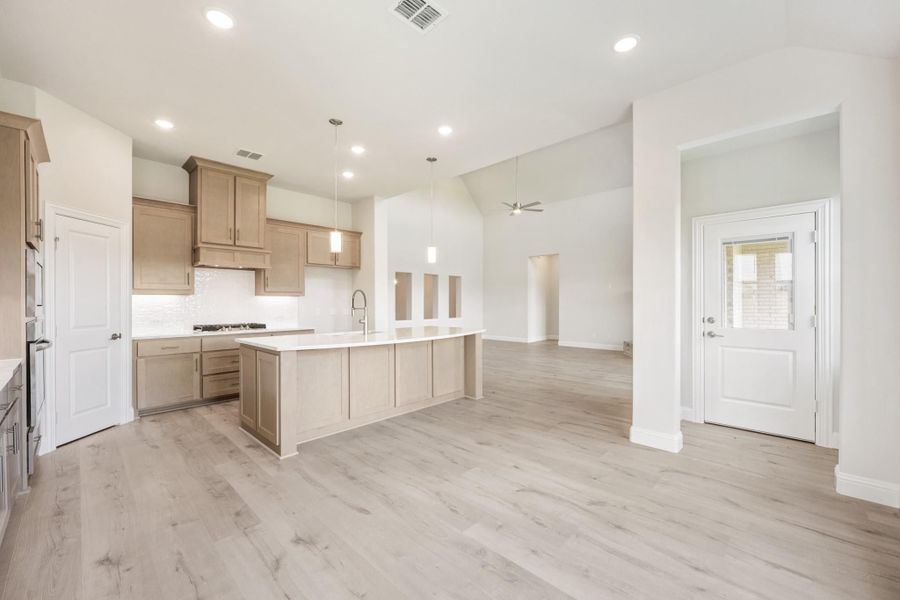 Open-concept kitchen with tan wood cabinets, large island, pendant lights, and light hardwood floors opening to living area