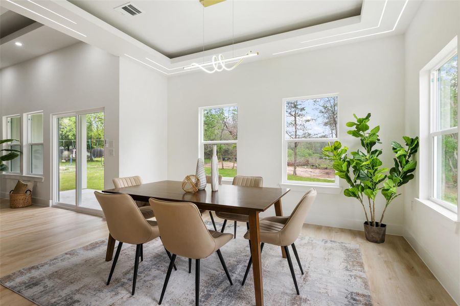Spacious dining area featuring a prominent tray ceiling with integrated linear lighting, light wood-finish flooring, and multiple windows providing natural light