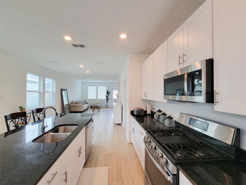 The kitchen features sleek black countertops, white cabinets, and stainless steel appliances. The space is bright with natural light from large windows, and there's a view of a cozy living room with contemporary furnishings.