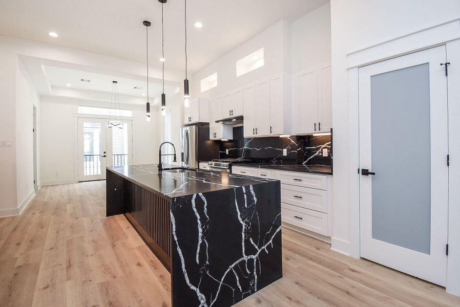 This modern kitchen features a sleek black marble island with matching countertops and backsplash, white cabinetry, stainless steel appliances, and elegant pendant lighting. Light wood flooring and a bright, open layout enhance the contemporary feel.