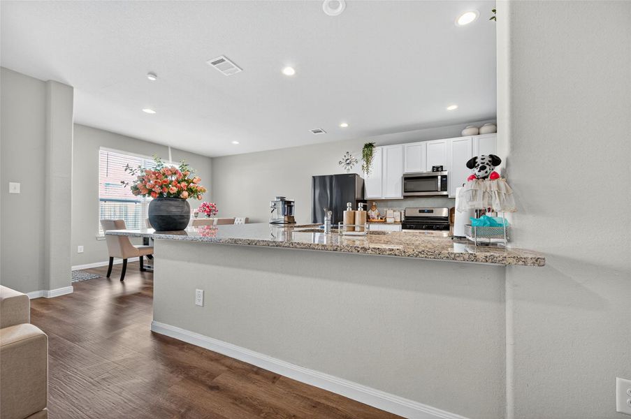 This photo showcases a modern kitchen and dining area with sleek granite countertops, stainless steel appliances, and white cabinetry. The room is well-lit with recessed lighting and features a cozy seating area by a window, accented with fresh flowers for a welcoming touch.