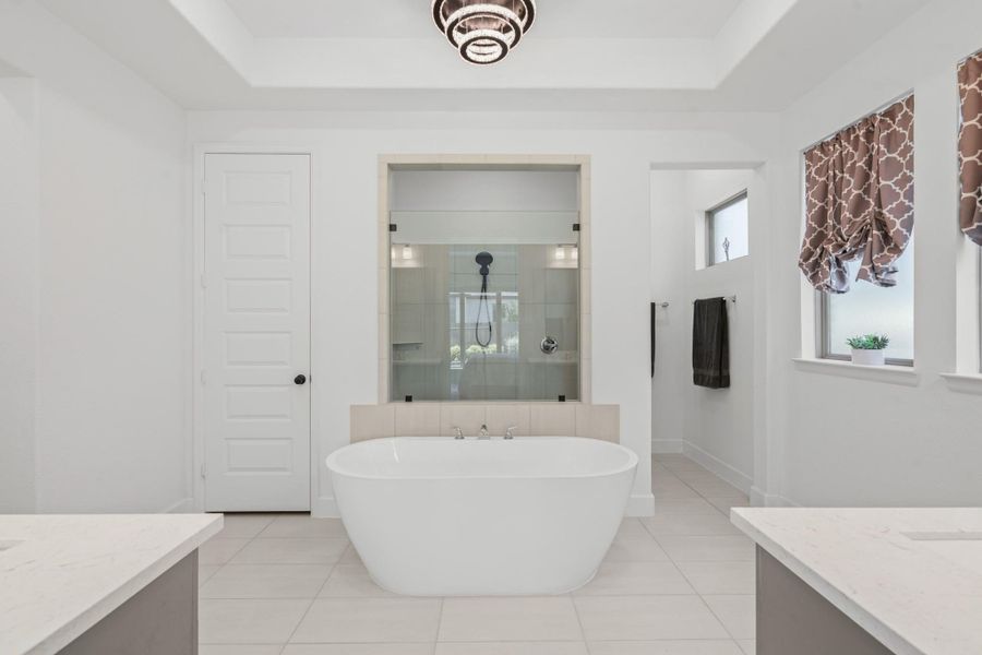 This elegant bathroom features a freestanding tub, a spacious glass-enclosed shower, and dual vanities. It is bright and airy with modern fixtures and patterned window treatments, creating a luxurious spa-like atmosphere.