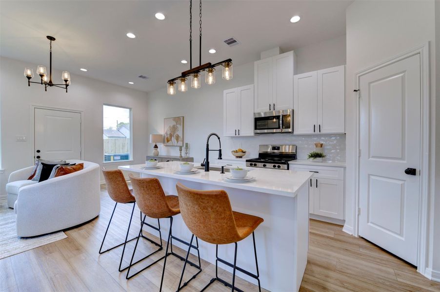 Kitchen island with quartz countertop, farmhouse sink, matte black coil-spring faucet, three leather bar stools on matte black frames, Edison rail pendant overhead, white shaker cabinets, and stainless gas range and microwave. Open to living room.
