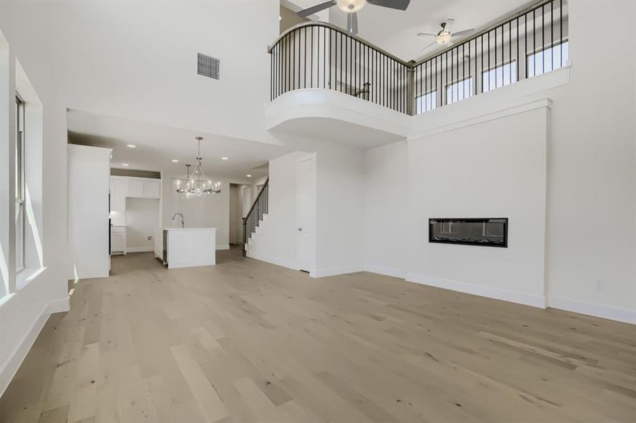Open-concept interior featuring light wood-finish flooring, a contemporary fireplace, a kitchen island with a sink and chandelier, a staircase with dark railings, and a second-story balcony with dark balusters