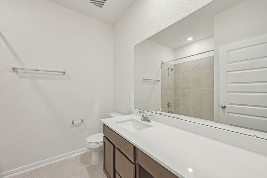 Secondary bath features decorative tile flooring, bath/shower combo with tile surround, stained wood cabinets, beautiful light countertops, mirror, sleek fixtures and modern finishes.