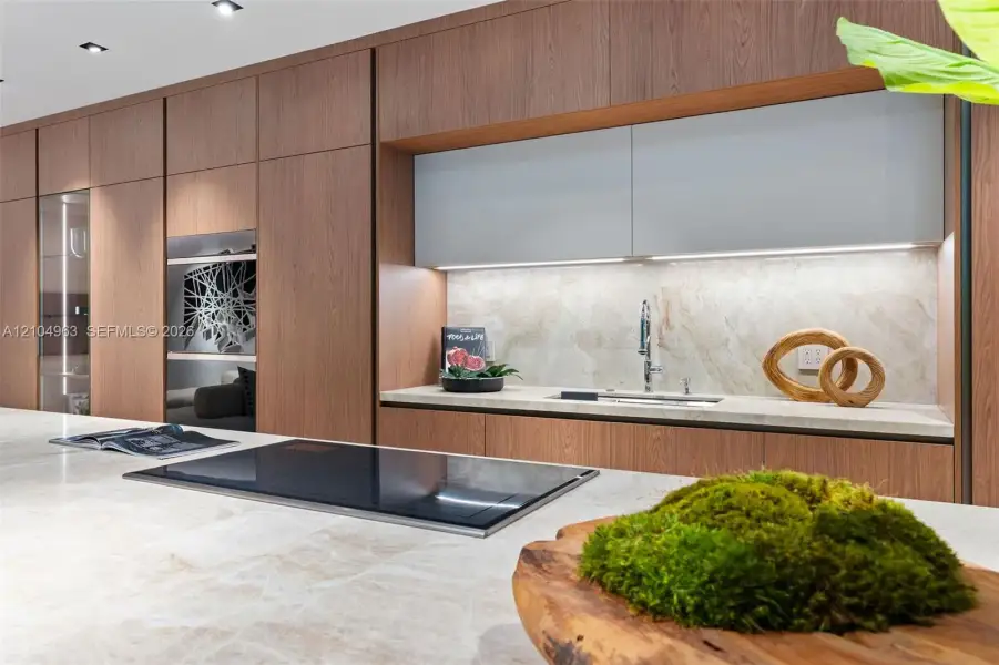 Modern kitchen featuring floor-to-ceiling wood-finish cabinetry, a large stone-surface island with an integrated induction cooktop, light-toned stone backsplash, and recessed ceiling lighting
