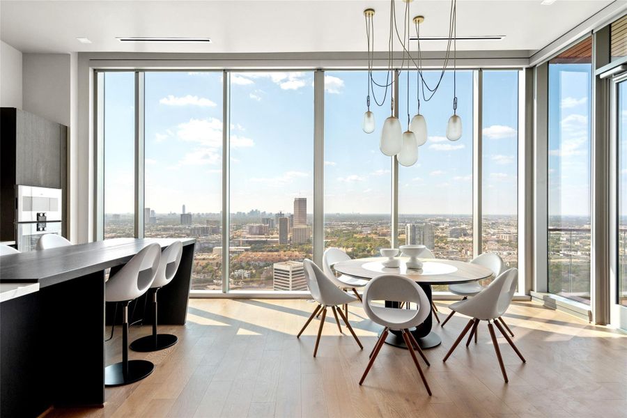 This is a modern, high-rise apartment featuring an open-plan dining area, floor-to-ceiling windows offering panoramic city views, and abundant natural light. The space is accented by stylish light fixtures.
