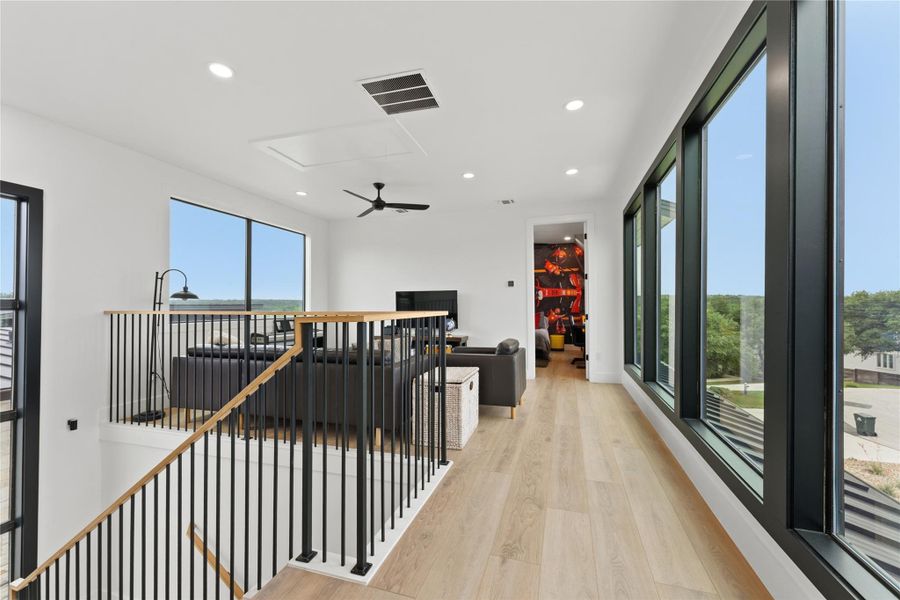 Upper-level hallway with full-height black-framed windows, light wood-finish flooring, recessed lighting, and a modern black ceiling fan Upper-level hallway with full-height black-framed windows, light wood-finish flooring, recessed lighting, and a modern black ceiling fan