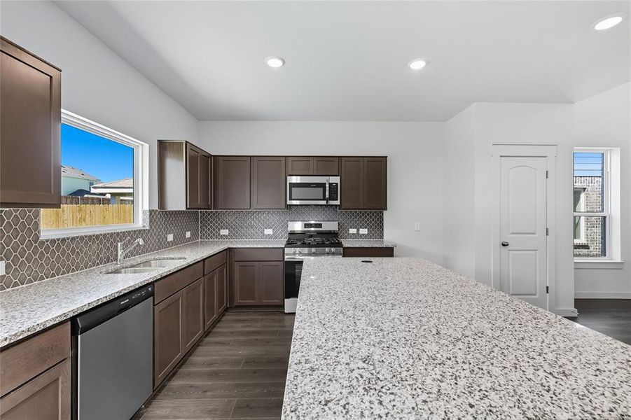 Kitchen with appliances with stainless steel finishes, dark wood-style floors, light stone countertops, backsplash, and dark brown cabinetry Kitchen with appliances with stainless steel finishes, dark wood-style floors, light stone countertops, backsplash, and dark brown cabinetry