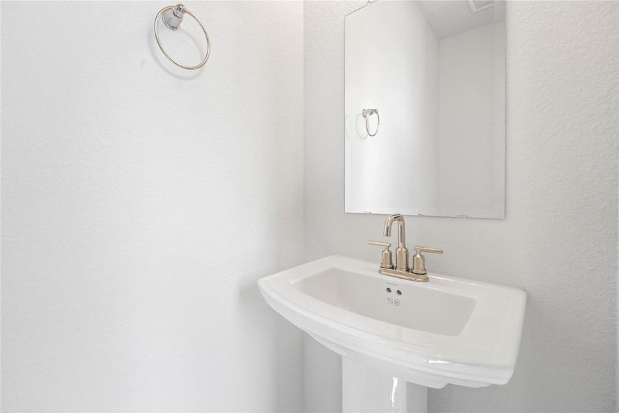 The first-floor powder room offers convenience and a modern aesthetic. It features a classic white pedestal sink complemented by a sleek, brushed-nickel faucet and a matching wall-mounted towel ring. A simple, large rectangular mirror enhances the sense of space and reflects the bright white walls, making this half-bath both functional and stylish.