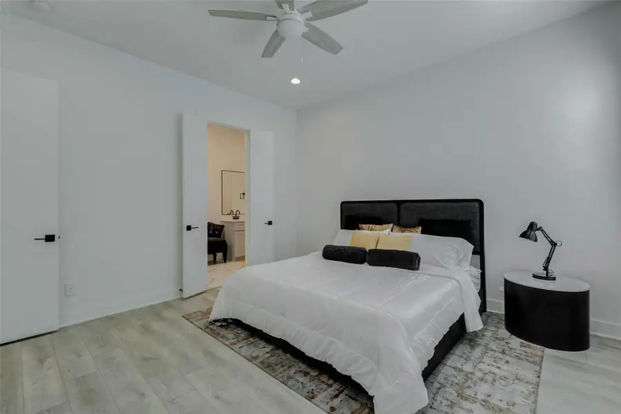 Spacious primary bedroom with soft neutral walls, light wood-look flooring, high ceiling, ceiling fan, and recessed lighting. The room offers a calm, airy atmosphere with a private connection to the en-suite bath, creating a comfortable retreat with clean finishes and a modern, move-in-ready feel.