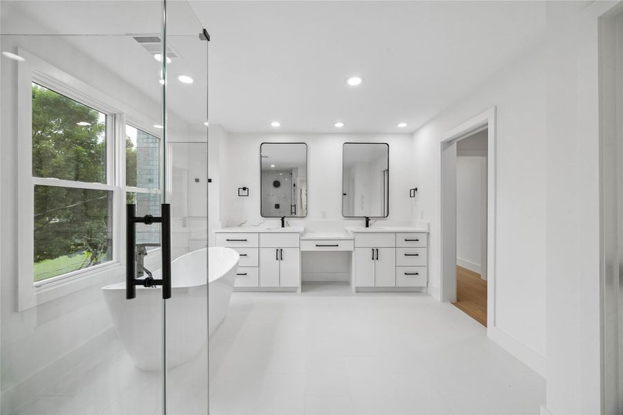 The primary bathroom combines clean contemporary finishes with a spacious layout centered around dual vanities, oversized mirrors, a freestanding soaking tub, and a large glass-enclosed shower. Oversized windows bring in natural light while maintaining a bright and refined spa-inspired setting throughout the space.