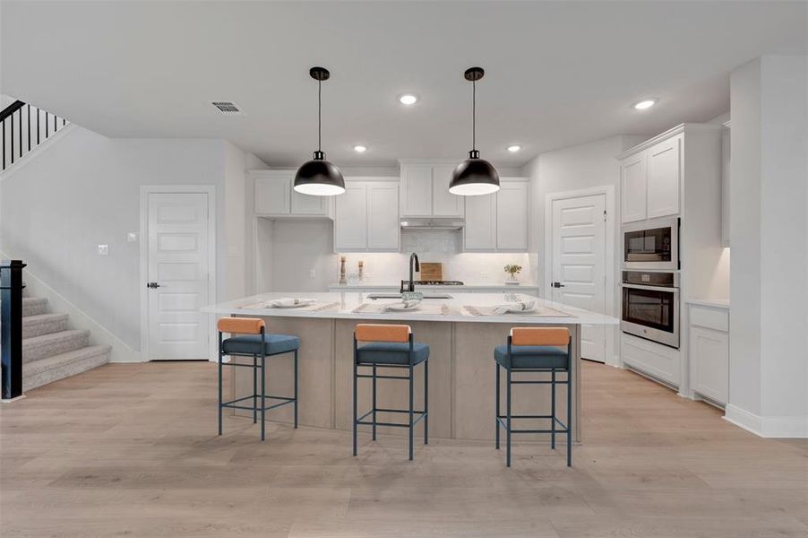 Open-concept kitchen featuring a large central island, light wood-finish flooring, white cabinetry, integrated stainless steel appliances, and two pendant light fixtures