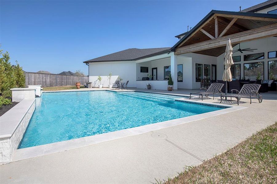 This home features a spacious backyard with a modern swimming pool, surrounded by a large patio area perfect for relaxing and entertaining. The house includes a covered outdoor seating area with ceiling fans, providing a comfortable and stylish setting.