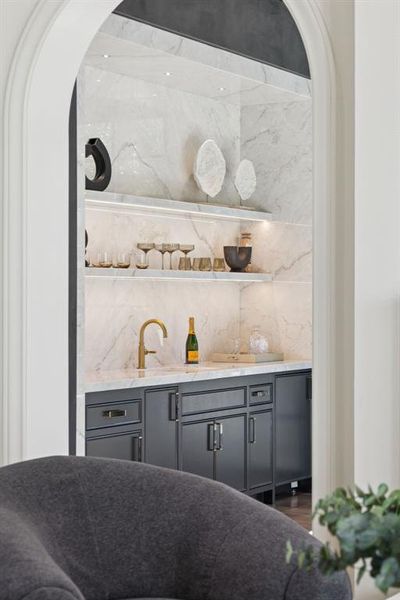 Indoor wet bar featuring gray cabinets, open shelves, arched walkways, and light stone countertops