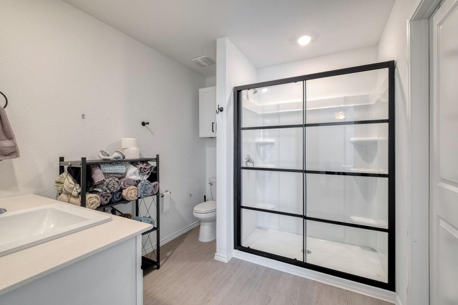 The bathroom features a spacious shower with black-framed glass doors, a white rectangular sink set into a light-colored countertop, and light wood-look flooring
