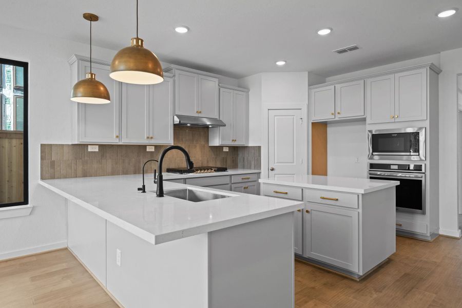 This beautiful kitchen features a bright, open layout with a large breakfast bar illuminated by elegant gold pendant lights that cast a warm, inviting glow. Sleek light grey cabinetry, natural wood flooring, and subtle metallic details create a perfect balance of modern luxury and timeless charm.