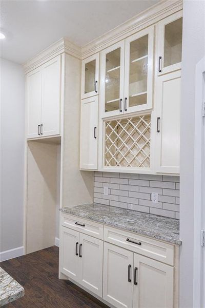 Bar area with decorative backsplash, light stone counters, glass insert cabinets, dark wood-style flooring, and white cabinetry Bar area with decorative backsplash, light stone counters, glass insert cabinets, dark wood-style flooring, and white cabinetry