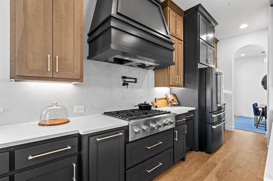 Kitchen featuring arched walkways, premium range hood, appliances with stainless steel finishes, backsplash, and light wood finished floors Kitchen featuring arched walkways, premium range hood, appliances with stainless steel finishes, backsplash, and light wood finished floors