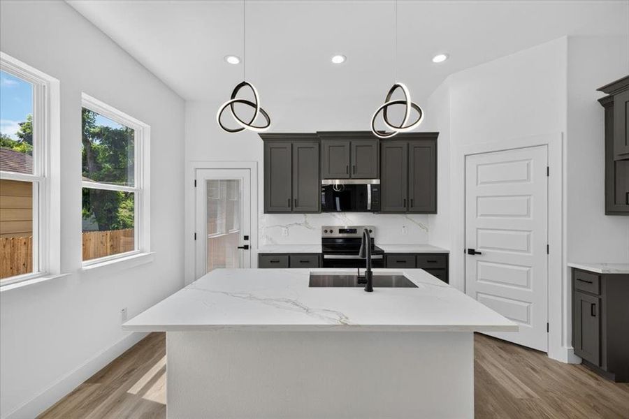 Kitchen featuring a central island with an integrated sink, quartz countertops, and dark gray cabinetry Kitchen featuring a central island with an integrated sink, quartz countertops, and dark gray cabinetry