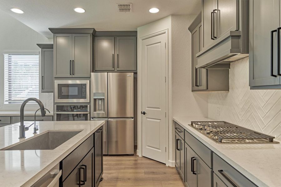 This modern kitchen features sleek gray cabinetry, stainless steel appliances, and a spacious island with a built-in sink. The light countertops and herringbone backsplash add a touch of elegance, while ample natural light enhances the inviting atmosphere.