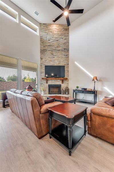 Living area featuring a stone fireplace, a high ceiling, light wood-style floors, and ceiling fan Living area featuring a stone fireplace, a high ceiling, light wood-style floors, and ceiling fan