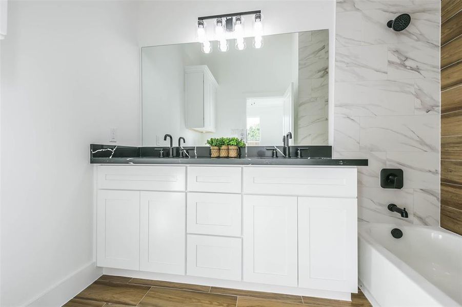 This is a modern bathroom featuring a sleek vanity with a black countertop and white cabinetry, a large mirror, and a tiled shower with a contemporary black shower head. The room is finished with wood-look flooring.