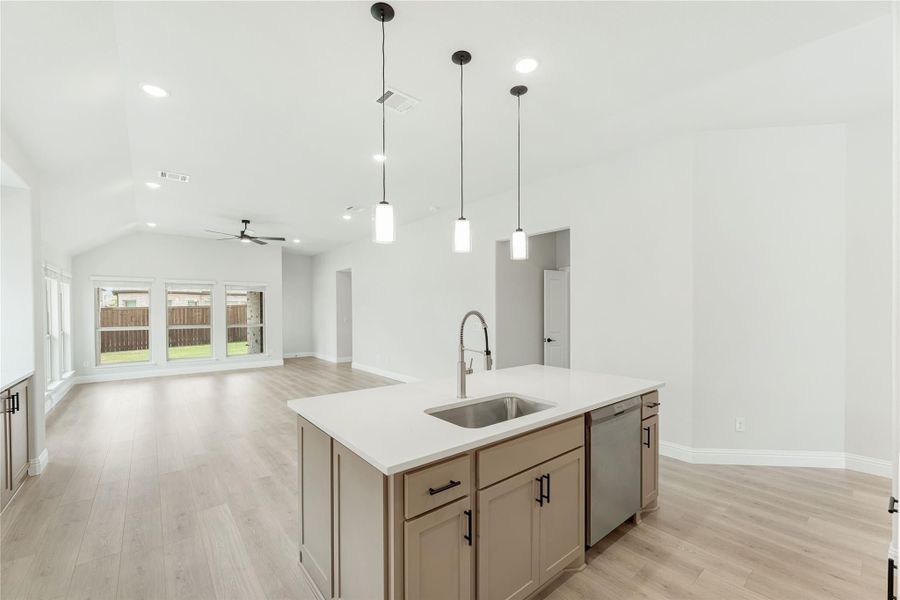Kitchen island with sink, pendant lights, and tan cabinets open to living area with vaulted ceiling and light wood floors
