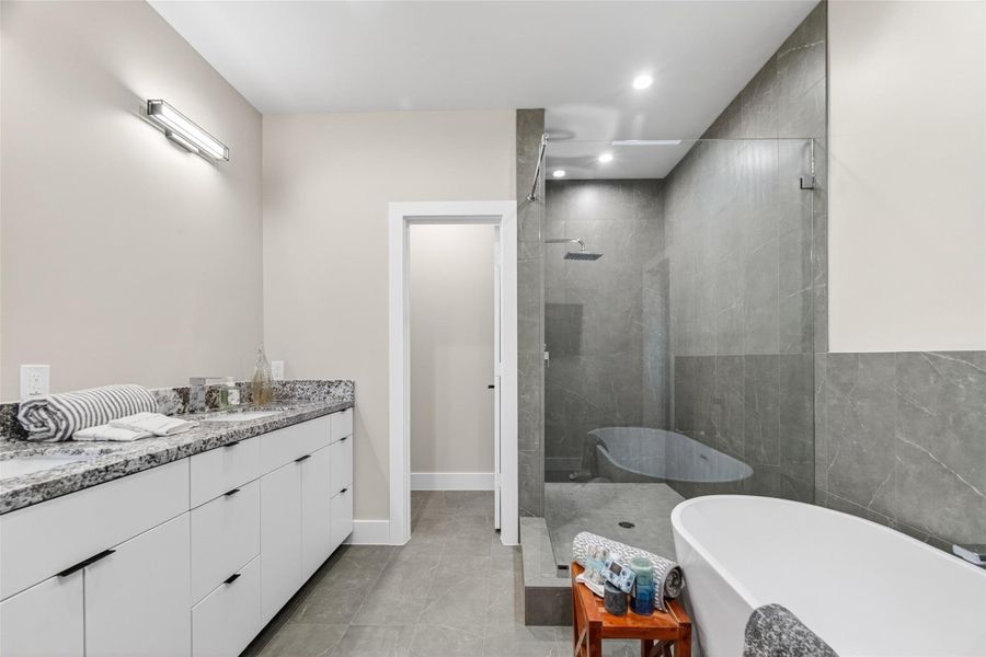The primary bath's double vanity stretches across the room with stone countertops, white cabinetry, and modern fixtures. The glass-enclosed walk-in shower, tiled floor to ceiling, is visible alongside the separate water closet, offering a fully functional spa-level layout.